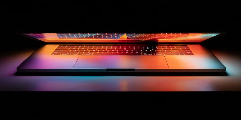 A laptop, partially open, displaying a vibrant, warm-toned graphic on its screen. The laptop keyboard is visible, and the surrounding area is dark.